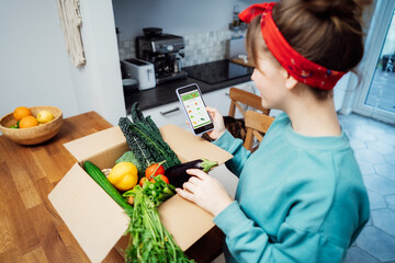Online home food delivery. Woman checking her online order list on her phone. Cardboard box with fresh vegetables and fruits standing on the kitchen table. Local farmer food. Start of a healthy life