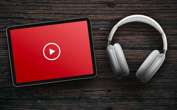 Play button on red screen of tablet and wireless headphones on dark wooden surface. Online streaming with white headset