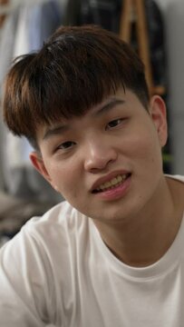 Vertical Screen: shoulder shot portrait of a shy asian young male looking at the camera and scratching head with a smiling face at a home bedroom background with daylight