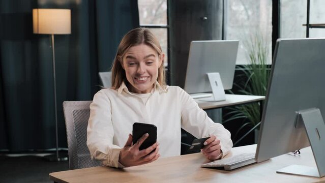 Happy millennial pretty woman holding bank card winning lottery, got amazing news, entering cvv code or payment information in mobile internet shopping application, feeling amazed of purchasing goods.
