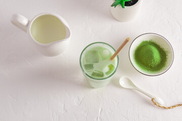 green healthy matcha latte tea with ice cubs on a white table with a bowl of matcha powder and milk jug with milk. top view. amino acids.