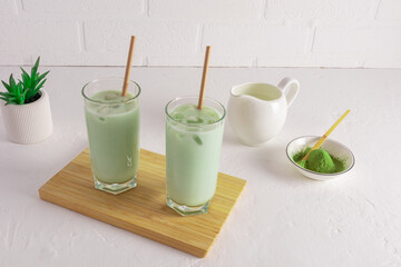 two glasses of freshly made matcha tea and milk on a wooden board on a white table against a brick white wall. an alternative to morning coffee.