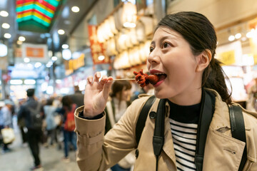 shoulder portrait of happy asian Japanese female tourist eating yummy grilled marinated baby octopus on busy teramachi street at nishiki market in Kyoto japan