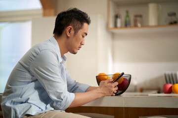 young asian man sitting at kitchen counter looking at cellphone