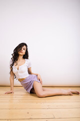 Fototapeta na wymiar A young brunette girl in a skirt and T-shirt with wavy hair is sitting on the floor on a white studio background.