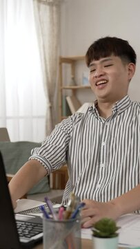 Vertical Screen: asian male employee closing his laptop and looking away into distance with a smile feeling happy about getting work done in the living room at home.