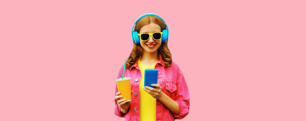 Portrait of stylish modern happy smiling young woman in headphones listening to music with smartphone wearing jacket on vivid pink background