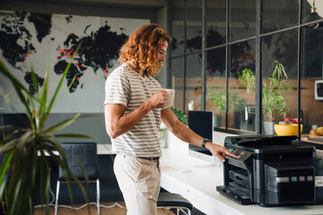 Young curly man drinking coffee and working with copy machine in office