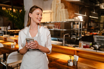 Young blonde woman wearing apron using cellphone while working in cafe