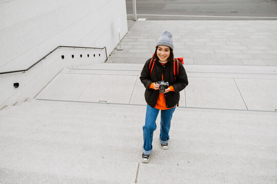 Cheerful asian woman taking pictures with vintage camera while climbing the stairs