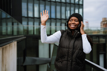 Cheerful afro woman waving hand while talking on mobile phone outdoors