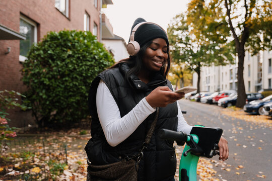 Cheerful afro woman using smartphone while standing with electric scooter outdoors