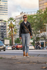 Adult 35s years old lesbian woman with sunglasses, leather jacket and blue jeans, walk along the street. Urban modern lifestyle in Barcelona city downtown in golden hour.