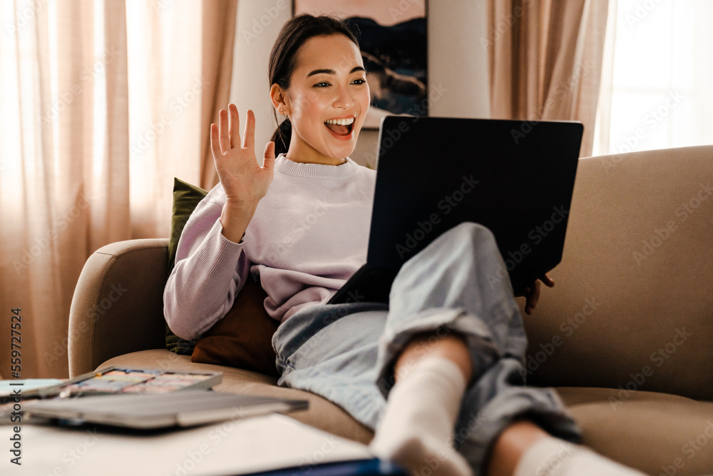 Wall mural Excited smiling asian woman making video call via laptop and waving hand while sitting on couch