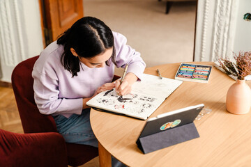 Fototapeta na wymiar Young woman artist drawing in sketchbook while sitting at table at home