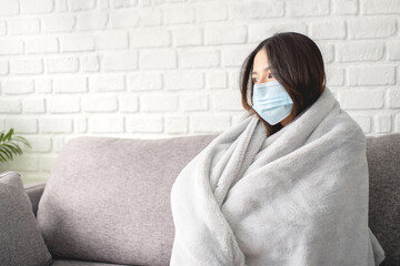 Young sick asian woman covered with blanket sits upset on sofa at home, wearing medical protective...