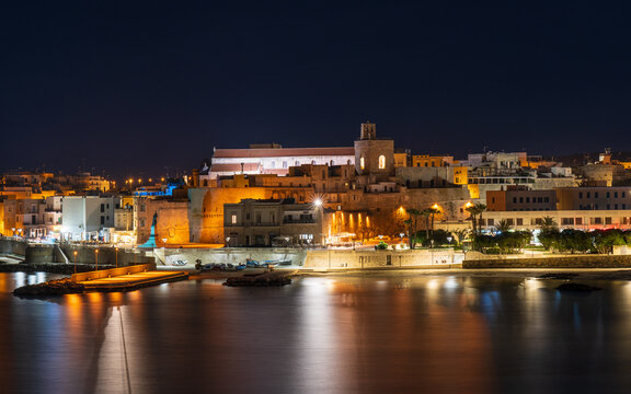 A night view of Otranto, a picturesque town on the Adriatic coast of Puglia, Italy. The lights of the buildings and the harbor are reflected in the calm water.