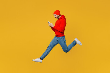 Fototapeta na wymiar Full body side view young caucasian man wear red hoody hat jump high hold in hand use mobile cell phone chat online isolated on plain yellow color background studio portrait. People lifestyle concept.