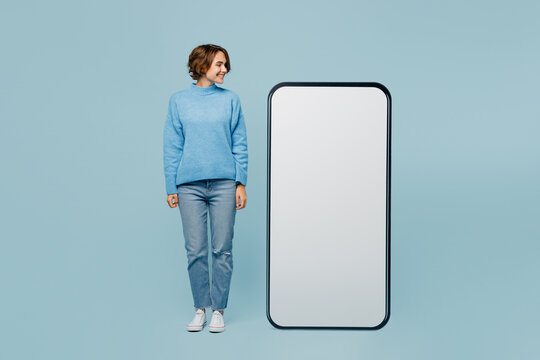 Full Body Happy Fun Cool Young Woman Wear Knitted Sweater Looking At Big Huge Blank Screen Mobile Cell Phone Smartphone With Workspace Mockup Area Isolated On Plain Pastel Light Blue Cyan Background.