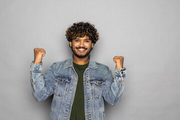 Young overjoyed happy fun Indian man doing winner gesture celebrate clenching fists say yes isolated on plain grey background studio portrait. People lifestyle portrait
