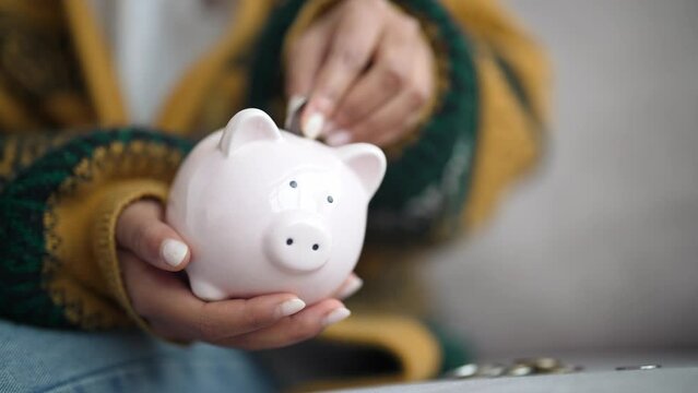 Close-up in hands girl holds piggy bank and puts coin money there. Cozy clothes and beautiful manicure. Home interior. Save up money investing in future life sitting on sofa in living room at home. 