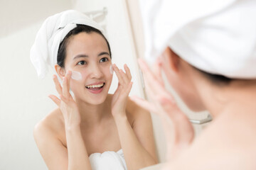 Happy young Asian woman applying face lotions while wearing a towel and touching her face in bathroom