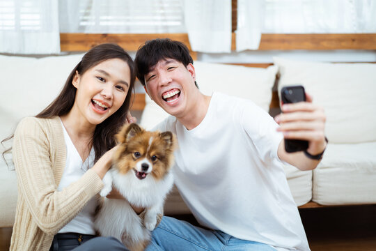 Happy cheerful Asian young couple taking a selfie photography or making a selfie video call on smartphone with their little dog in living room. Man and woman smiling to camera on smartphone.