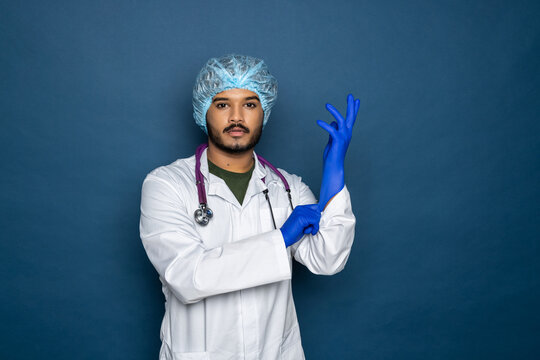 Indian Doctor Or Physician Male Looking At Hand With Focused Expression As Putting On Blue Surgical Latex Glove Isolated On Blue Background