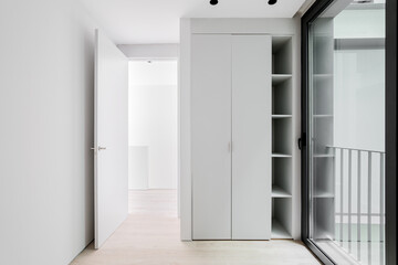 Small entrance hall for meeting guests with large glass front door in black metal frame, which reflects part of railing on street. White wooden wardrobe with closed doors and shelves for small items.