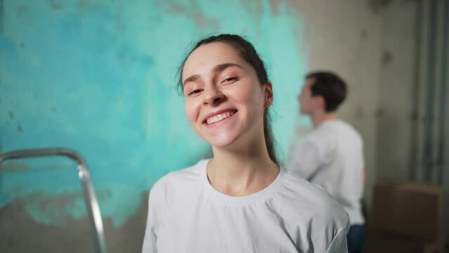 Portrait girl house painter stands smile, looking at camera. Man working, painting wall paint roller with primer or blue paint. Wallpaper for painting. Interior, renovation. Professional occupation.