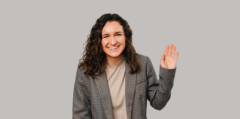 Photo of charming happy young woman in casual suit making Hello gesture over grey background. © Vulp