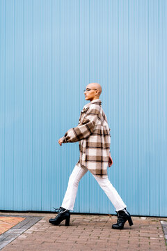 Woman with shaved head walking on footpath by blue wall