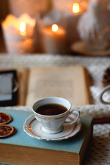 Cup of tea, books, tablet, bowl of cookies, various spices, pine cones and lit candles. Hygge at home. Selective focus.