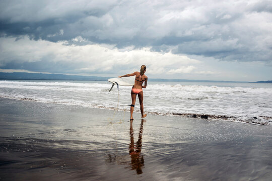 Rear view of woman carrying surfboard at beach against cloudy sky