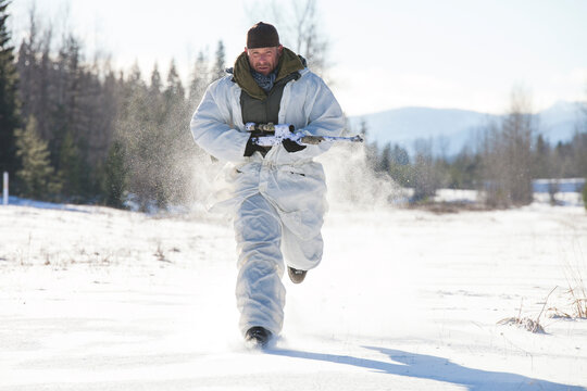 A hunter wearing  a cold-weather camouflage outfit carries his rifel while running through a snow-covered field.