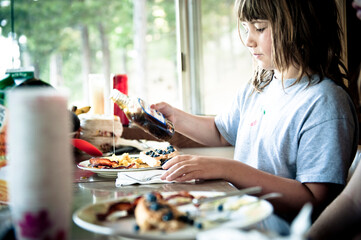 A young girl pours syrup on her breakfast: bacon, eggs and pancakes.