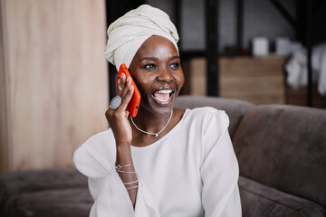 Excited African woman in white turban, white dress sitting on cozy couch talks by phone home opens...