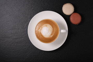 White cup with cappuccino on a dark background flat lay. breakfast on a black background. Coffee with macaroon on a black background.