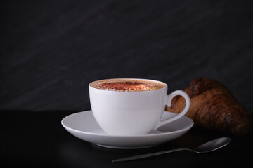 Cappuccino with croissant on a black background. White cup with cappuccino on a dark background. Italian breakfast on a black background.