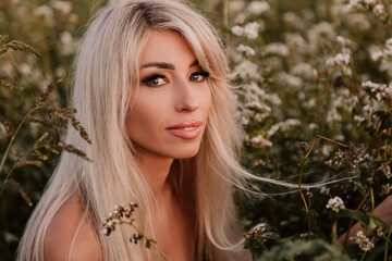 Naklejka premium Blonde naked woman posing in the big endless field of daisies in summer evening. Lady wear pink dressed, looking at camera smiling, touching the plant green blossoming flower. Moody atmosphere 