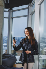 Young smart businesswoman in casual clothes working with phone while standing near window and reporting and writing notes and against blurred modern office interior.