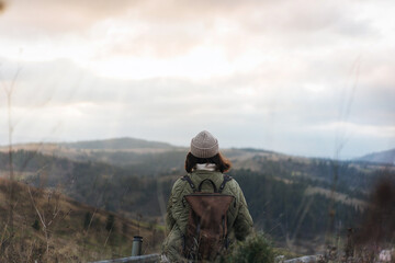 View from the back of hiker girl wearing backpack and beanie hat sitting on edge of cliff and enjoying landscape, sunset, mountains and clouds.