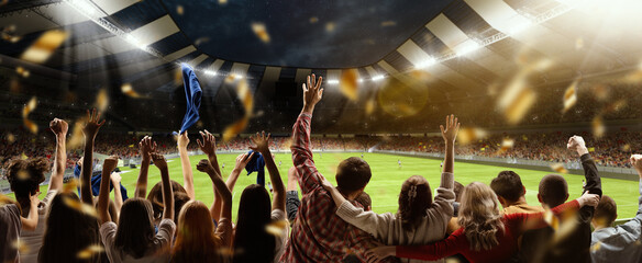 Back view of football, soccer fans cheering their team, holding flag at crowded stadium at evening...