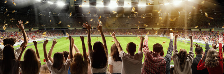 Back view of football, soccer fans emotionally cheering their team at crowded stadium at evening...