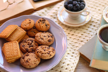 Plate of cookies, cup of tea, fresh blueberries, dry oranges, stack of books, reading glasses and tablet on the table. Hygge at home. Selective focus.