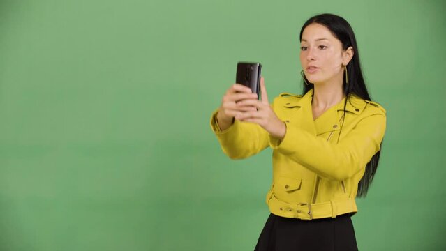 A young beautiful Caucasian woman takes selfies with a smartphone - green screen studio