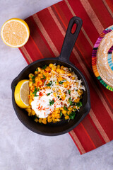 Esquites, grilled corn, served with grated cheese, sour cream, chili powder and slice of lemon in cast iron pan. Mexican street food meal. Top view