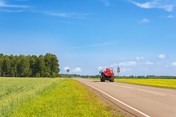 Red fuel truck on an empty rural road among golden flowering farm fields with rapeseed. Summer landscape. Fuel truck is driving along a road. Lorry with orange tanker. Back view.