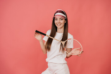 Satisfied European young woman with loose hair in white t-shirt and transparent visor, smiles looks at camera, holds badminton racket against studio pink background. Sport , healthy lifestyle concept.