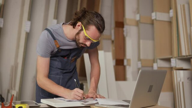 Male carpenter working with computer laptop in workshop. craftsman wearing safety glasses checking order of clients or learning online. tired Business Owner wood worker In Workshop.